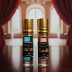 Two perfume bottles with gold caps on a wooden surface, with a blurred background of red curtains and a chandelier.